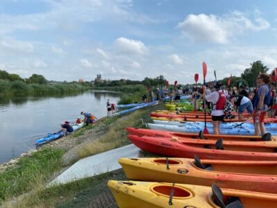 Zdjęcie przedstawia kajaki, uczestników i rzekę, zostało zrobione podczas II Akademickiego Spływu Kajakowego