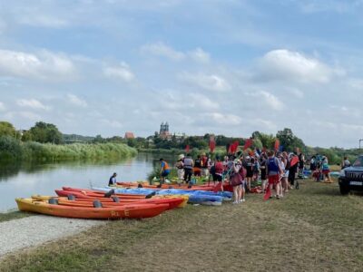 Zdjęcie przedstawia kajaki, uczestników i rzekę, zostało zrobione podczas II Akademickiego Spływu Kajakowego