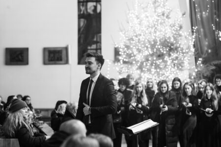 Na zdjęciu widać chór i dyrygenta stojącego przed nim tuż po wykonaniu utworu. zdjęcie jest czarno-białe, wykonane w kościele na tle choinki z lampkami.