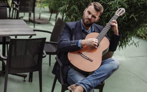 Na zdjęciu Łukasz Kuropaczewski siedzi przy stoliku z gitarę w reku