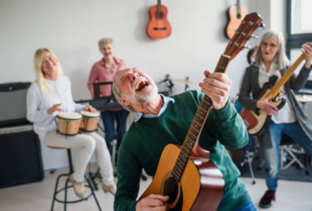 Kkolorowa fotografia przedstawia na piewrwszym planie starszego mężczyznę z gitarą, który śpiewa utwów, w tle widać innych ludzi, kobieta trzyma na kolanach bębenki